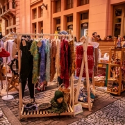 Imagem de uma feira de rua. Nela uma arara om roupas e uma mulher olhando as roupas. Outras pessoas transitando no local.