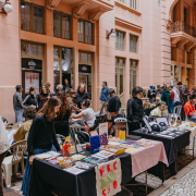 Fotografia retirada na travessa dos cataventos. Na imagem, 7 mesas com produtos de arte gr&aacute;fica como desenhos, pinturas em tecidos, entre outros itens. Em torno das mesas, os vendedores, artistas, e o p&uacute;blico visitante. Em torno de 30 pessoas na foto que tem como fundo, a Casa de Cultura Mario Quintana. 